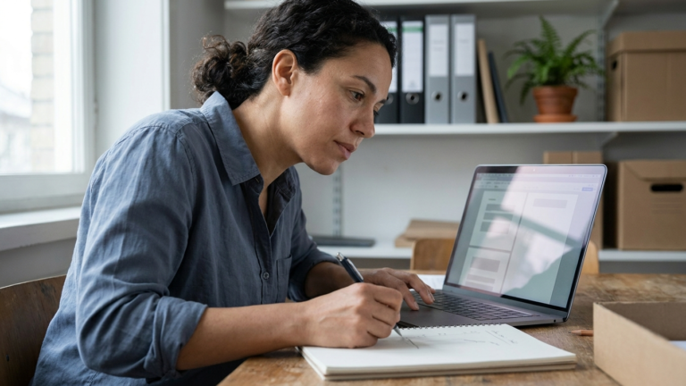Mulher consultando um notebook enquanto faz anotações em um caderno, em um escritório.